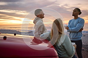 Group Of Friends Standing Chatting By Car With Hot Drink At Beach Watching Sunrise Together photo