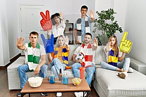 Group of friends supporting football team watching tv al home sitting on the sofa with open hand doing stop sign with serious and