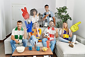 Group of friends supporting football team watching tv al home sitting on the sofa pointing thumb up to the side smiling happy with