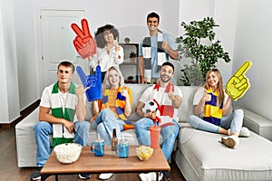 Group of friends supporting football team watching tv al home sitting on the sofa smiling happy and positive, thumb up doing