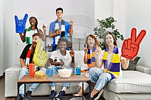 Group of friends supporting football team watching tv al home sitting on the sofa surprised with an idea or question pointing