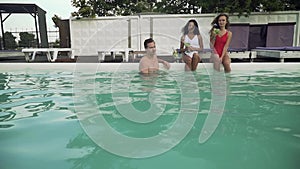 A group of friends vacationing lounging by the outdoor pool. Guy standing in a swimming pool and two beautiful