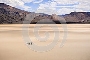 A group of friends walking across the surface of the Racetrack Playa on a sunny day; mountains and white clouds in the background