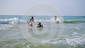 Group of friends young men, women in swimwear run into sea waves, fall, splash in waves at tropical travel. Back view