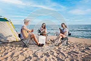 Group of happy Asian friends playing guitar and singing with clap while picnic and camping on the beach in outdoors vacation