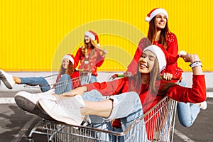 Group of happy cheerful young women in christmas sweaters and santa claus hats walking down city street riding shopping trolleys