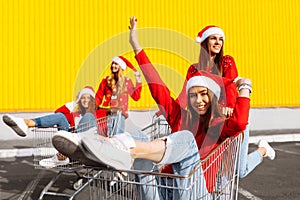 Group of happy cheerful young women in christmas sweaters and santa claus hats walking down city street riding shopping trolleys