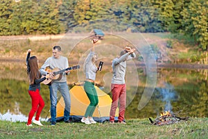 Group of happy friends with guitar, having fun outdoor, dancing and jumping near the lake in the park background the Beautiful sky