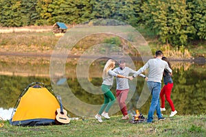 Group of happy friends with guitar, having fun outdoor, dancing and jumping near the lake in the park background the Beautiful sky