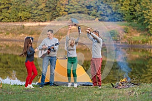 Group of happy friends with guitar, having fun outdoor, dancing and jumping near the lake in the park background the Beautiful sky