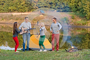 Group of happy friends with guitar, having fun outdoor, dancing and jumping near the lake in the park background the Beautiful sky