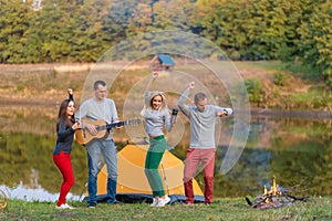 Group of happy friends with guitar, having fun outdoor, dancing and jumping near the lake in the park background the Beautiful sky