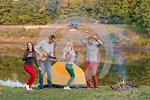 Group of happy friends with guitar, having fun outdoor, dancing and jumping near the lake in the park background the Beautiful sky