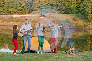 Group of happy friends with guitar, having fun outdoor, dancing and jumping near the lake in the park background the Beautiful sky