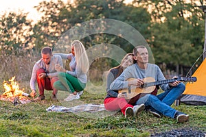 Group of happy friends with guitar, having fun outdoor, near bonfire and tourist tent. Camping fun happy family