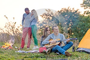 Group of happy friends with guitar, having fun outdoor, near bonfire and tourist tent. Camping fun happy family