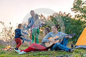 Group of happy friends with guitar, having fun outdoor, near bonfire and tourist tent. Camping fun happy family