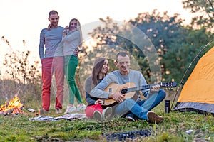 Group of happy friends with guitar, having fun outdoor, near bonfire and tourist tent. Camping fun happy family