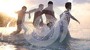 Group of happy friends at sunset running into water getting wet and making splashes swimming in the sea