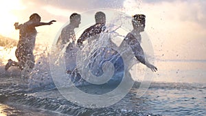 Group of happy friends at sunset running into water getting wet and making splashes swimming in the sea