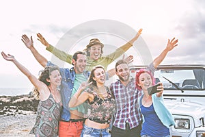 Group of happy friends taking a selfie using mobile smart phone during a road trip with jeep car - Travel people having fun