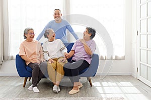 The group of happy and healthy three Asian senior women sits together on a sofa with an Asian senior man standing in the back.