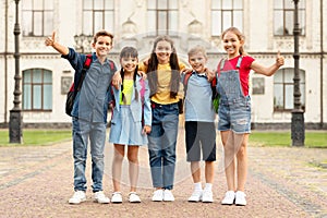 Group of happy multiethnic kids with backpacks posing outdoors, showing thumbs up