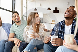 Group of happy multiracial friends drinking coffee in living room, having conversation, enjoying time together