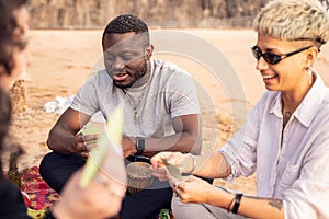 Group of happy multiracial friends having fun together on the beach, playing cards on picnic, laughing. Mixed race people