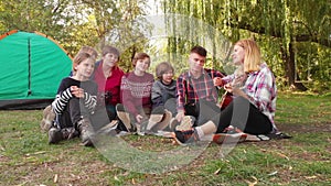 Group of happy people with children sing while relaxing on summer vacation on camping trip. Woman playing acoustic