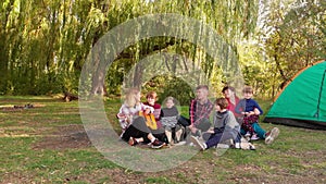 Group of happy people with children sing while relaxing on summer vacation on camping trip. Woman playing acoustic
