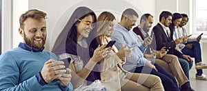 Group of happy young men and women sitting in row and text messaging on their cellphones