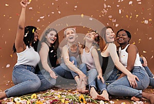 Group of happy women sitting on the brown background while petals from flowers falling. Laughing females of different races and