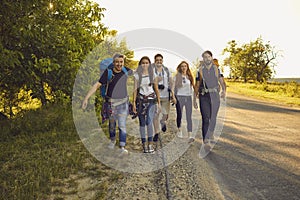 Group of happy young friends with backpacks walking along road in countryside. Hikers on tourist trip in summertime