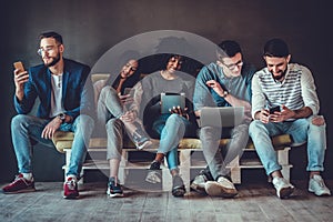 Group of happy young people sitting on sofa and using digital tablet and laptop