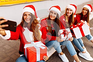 Group of happy young women in santa claus hats holding gift boxes and taking selfie on mobile phone while sitting on sidewalk