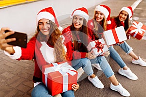 Group of happy young women in santa claus hats holding gift boxes and taking selfie on mobile phone while sitting on sidewalk