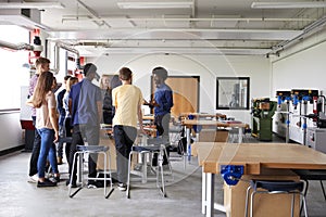 Group Of High School Students Standing Around Work Bench Listening To Teacher In Design And Technology Lesson