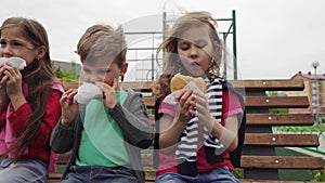 Group of hungry kids sitting on bench and eating burger outdoor on children playground. Slide shot. The lockdown and