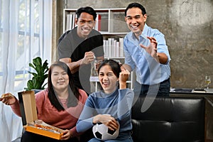 Group of joyful Asian friends sit on sofa watching and cheering football team on TV together