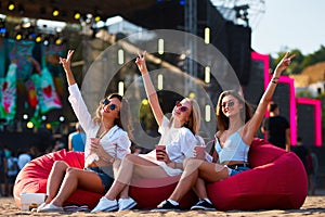 Group of joyful girls on red bean bags at sunset, cheering with drinks, enjoying live music at sandy beach fest. Casual
