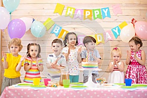 Group of kids clap their hands near birthday cake, balloons on background