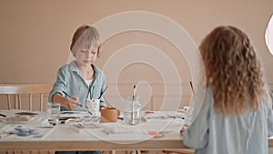 Group of kids and their teacher leaning over table with watercolor paints and painting self-made clay items. Children