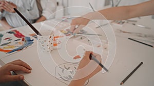 Group of kids and their teacher leaning over table with watercolor paints and painting self-made clay items. Children