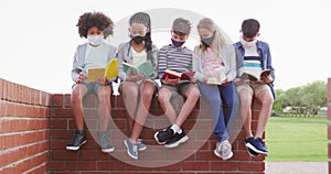 Group of kids wearing face masks reading books while sitting on a brick wall