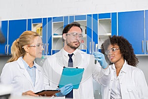 Group of Laboratory Scientists Examining Green Liquid In Test Tube Using Tablet Computers, Mix Race Team Of Lab Workers