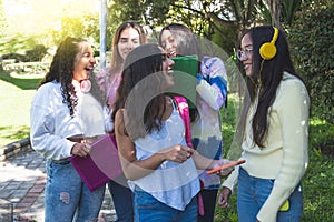 Group of Latinx teenage friends review their homework and laugh with their headphones and smartphones walking in the middle of a photo