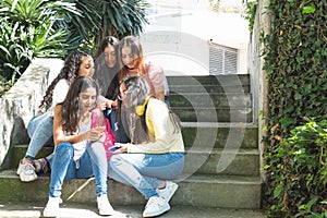 Group of Latinx teenage girls students interacting with their cell phones sitting in some bleachers outdoors photo