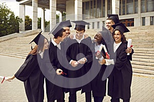 Group of laughing happy multiracial graduates hugging outdoors holding diploma.