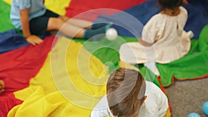 Group of little children playing around on rainbow play parachute and throwing plastic balls. Multiracial nursery